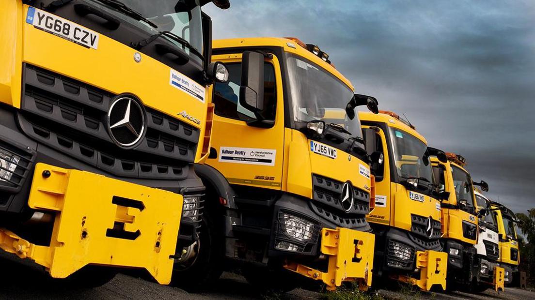 A row of yellow gritter lorries. Several front cabs can be seen with metal attachments on their front to become ploughs. Overhead are overcast skies.