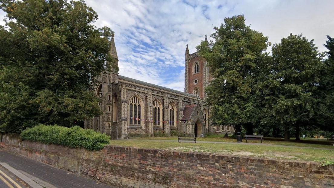 External view of St Peter's Church with its brick built tower. A number of large mature trees can be seen in the churchyard. The edge of the church has a brick wall, which drops down to St Peters Road in the foreground. 