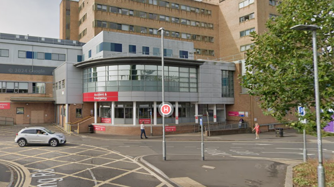 The entrance of the A&E department at Yeovil District Hospital. The entrance is rounded and is made up of a mixture of grey cladding and glass. The wider hospital building behind it is brown and has at least six storeys.