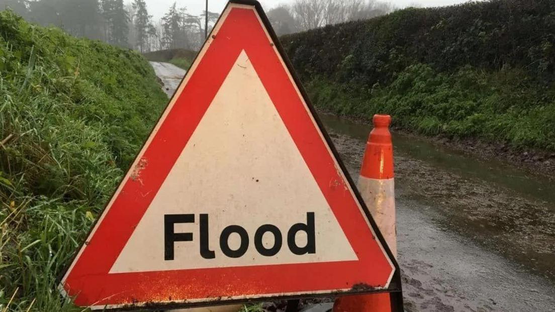 A "flood" road warning sign on a single track road with grass verges either side of the road which has a lot of rainwater on it. An orange and white traffic cone is next to the sign.