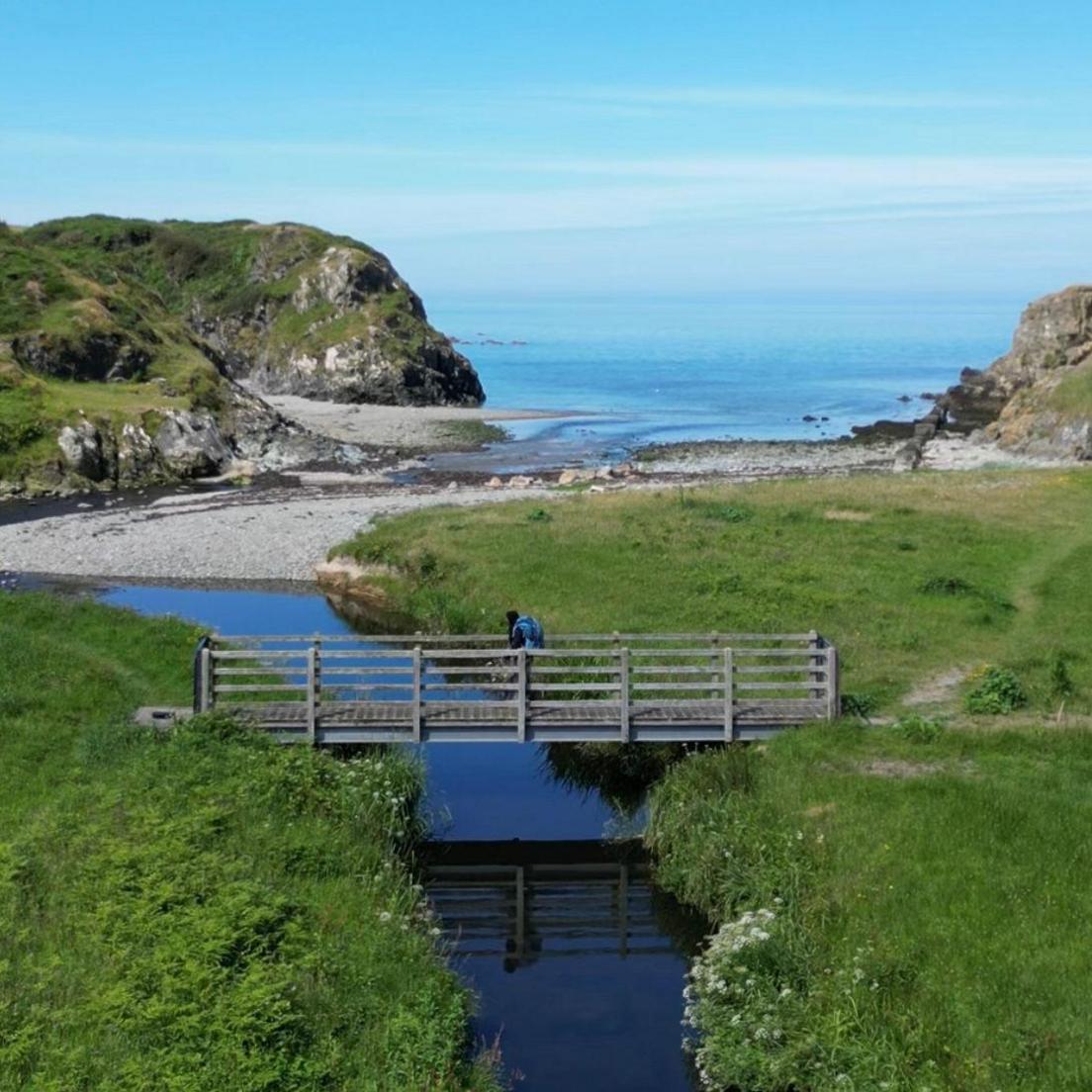 Amina is walking over a bridge by the coast. She has a backpack on. There is a lot of scenery behind her, including green hills and a blue sea. 