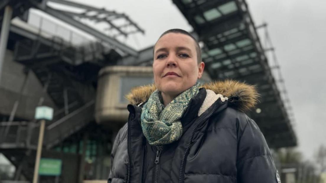 Campaigner Jo Howell stands in front of the National Glass Centre on the banks of the River Wear, wearing a thick waterproof jacket and a scarf. She has short, dark hair. 