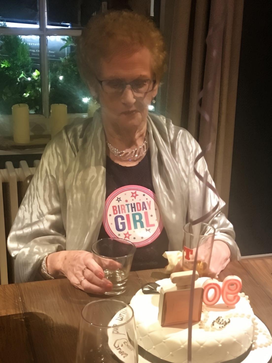 Ellen Watson, wearing a silver draped jacket over a black top with dark framed glasses and a silver necklace, sits at a restaurant table in front of a birthday cake with "90" candle and three glasses.  She is wearing a large badge saying "birthday girl". There is a balloon ribbon in the foreground and candles on a window sill behind her. 