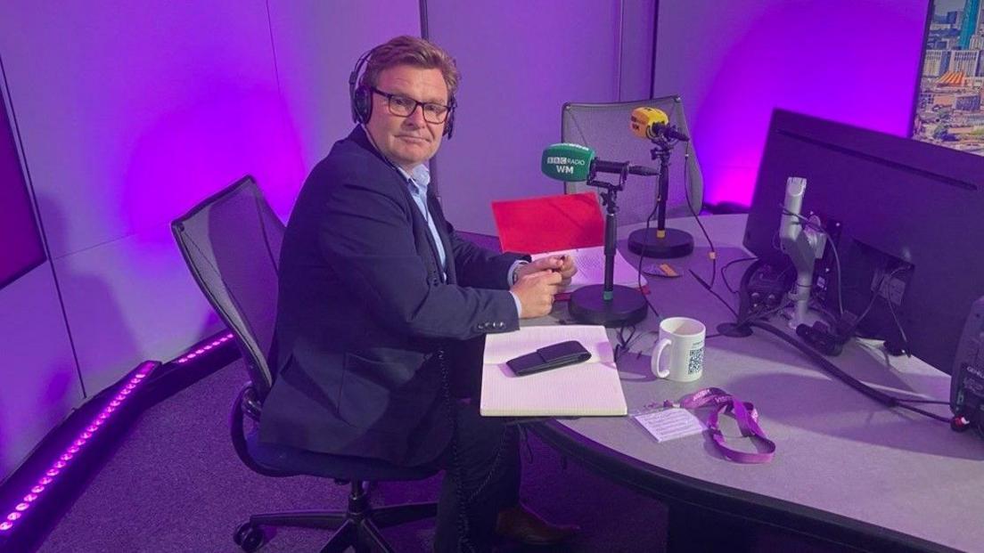 A man in a shirt and blue jacket is sitting side on to the camera, but with his head facing the camera. He is sitting by a green microphone in a radio studio that has a purple background.