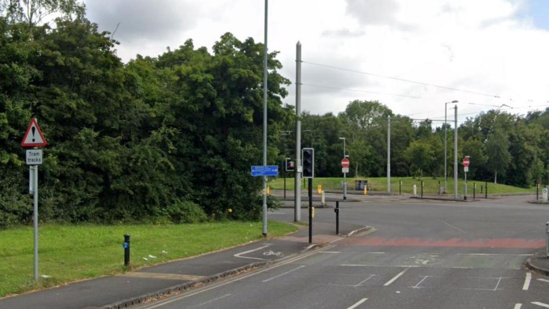 A road just before a major junction showing a traffic crossing, bicycle lanes and a pavement along with a warning sign for tram tracks. Toward the left of the road, a green verge can be seen with trees and shrubs. 