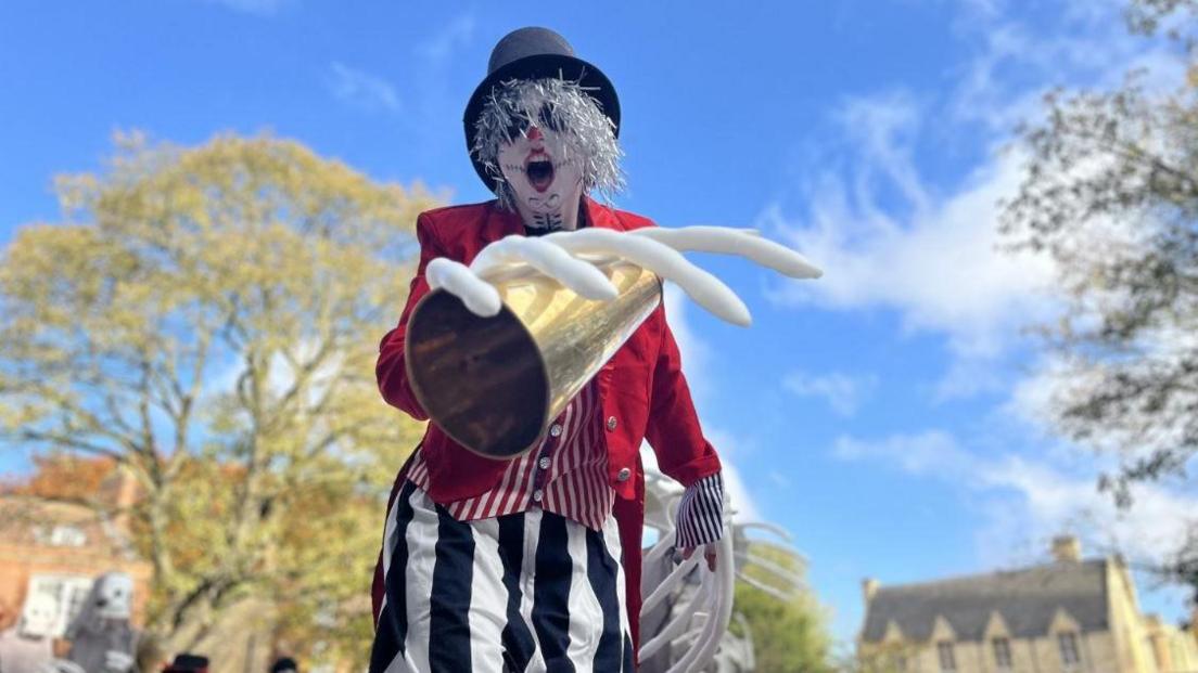 A person dressed as a skeleton with a black hat on, a silver wig and dressed in a red and white striped waistcoat, a red jacket and black and white striped trousers. They have fake skeleton hands on and are looking directly into the screen with their mouth open. They have skeleton face paint on. 