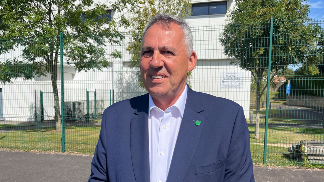 Constable Andy Jehan, in a blue suit and white shirt, with a green badge on his lapel, standing in front of a fence at Oakfield.