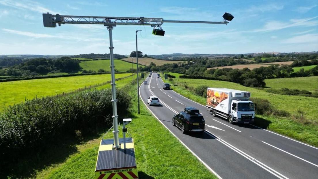 Cameras are mounted on a metal post overlooking a single carriageway road which is leading through undulating hills and fields.