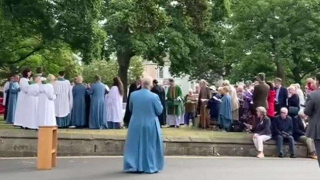 Worshippers gathered outside on the lawn, under the trees.  The Church officials are in their garments standing in the foreground and the congregation are spread out, with some sat on a nearby wall.