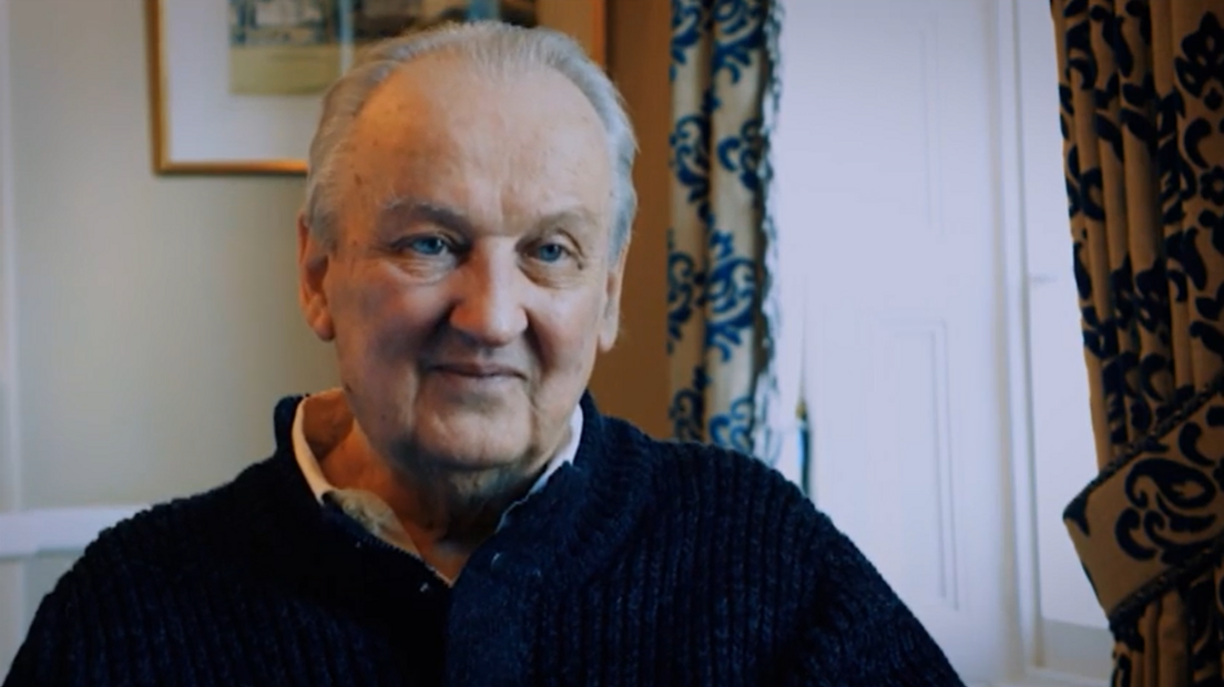 Douglas Gowan wearing a dark knitted jumper sits indoors beside a bright window. Patterned curtains frame the window, while a framed picture hangs on the wall behind. The photograph is warmly lit and focuses closely on his face and upper body.