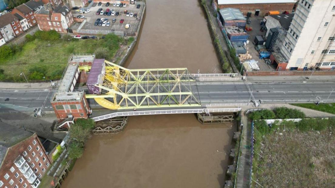 A drone shot of a yellow steel bridge over a river. There are city centre buildings and wasteland next to the river.