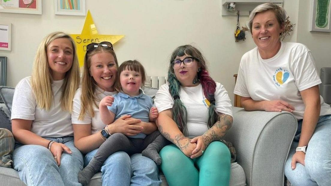 Four women sit in a row on a sofa, with a four-year-old on one of their laps, all smile to the camera.