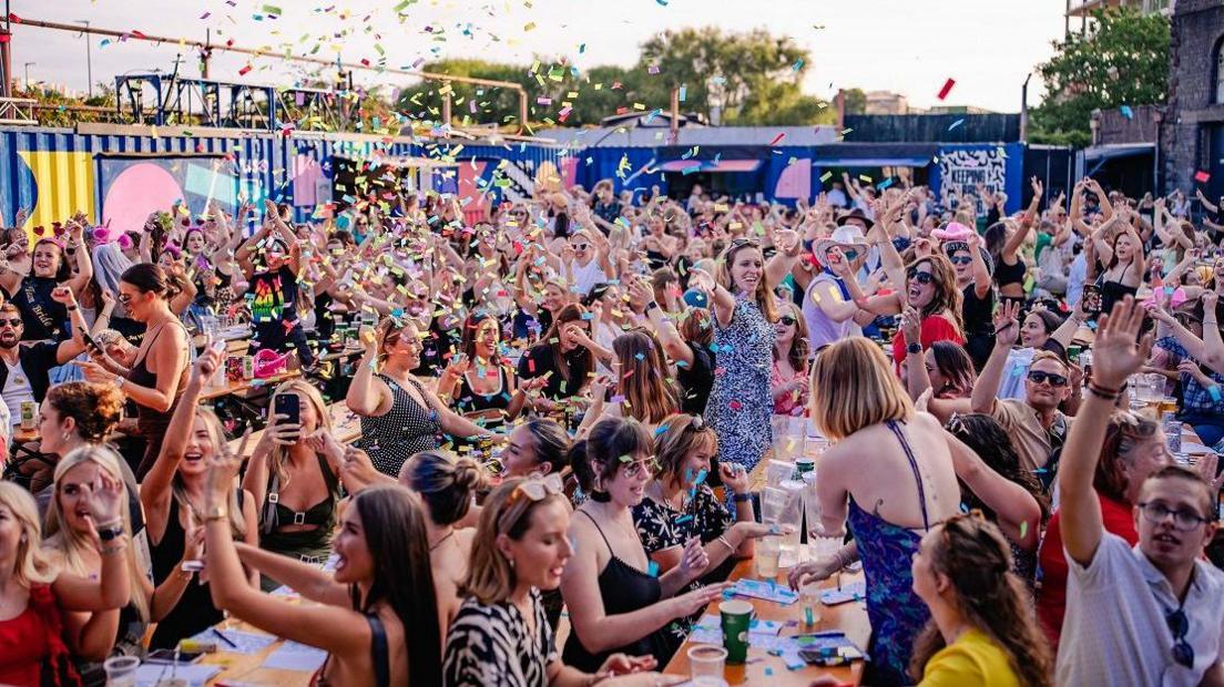 A crowd of people sit at tables outside a court yard on a bright day. People raise their hands as confetti falls from the sky