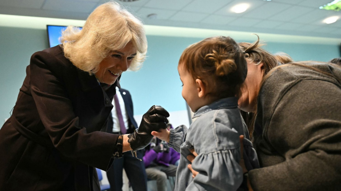 Queen Camilla interacts with a young child in a hospital setting, reaching out to hold the child's hand as it is supported by its parent. The Queen is wearing a black leather glove