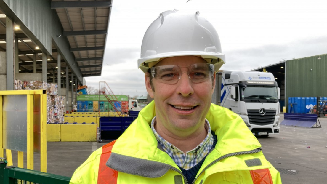 A close-up of councillor Richard Wilkins wearing a high vis jacket and white hard hat, smiling to camera with the recycling centre in the background and a lorry