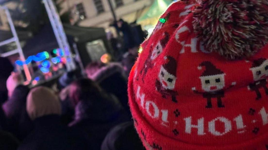 Crowds of people slightly blurred at a lights turn on, with a child's head, with a red bobble hat saying, Ho Ho Ho, and a Santa on looking at a stage