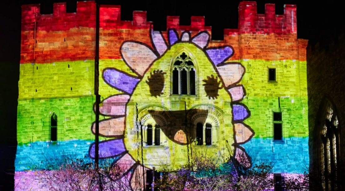 Colour the Castle projection on Durham castle by Dutch production house Mr Beam during Lumiere in 2023. It features a smiling flower with pink, purple and peachy petals against a background of purple, blue, green, yellow, orange and read streaks.