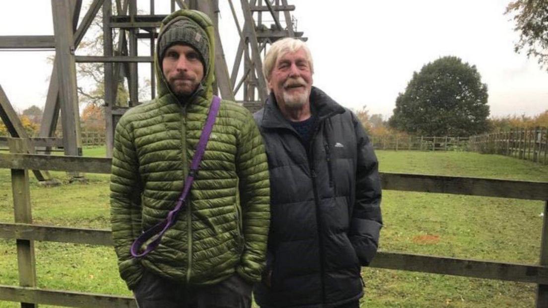 James Rogers and Alan Rogers stood in front of the wooden Brinsley Headstocks. James is in a green coat, while Alan is in a black coat.