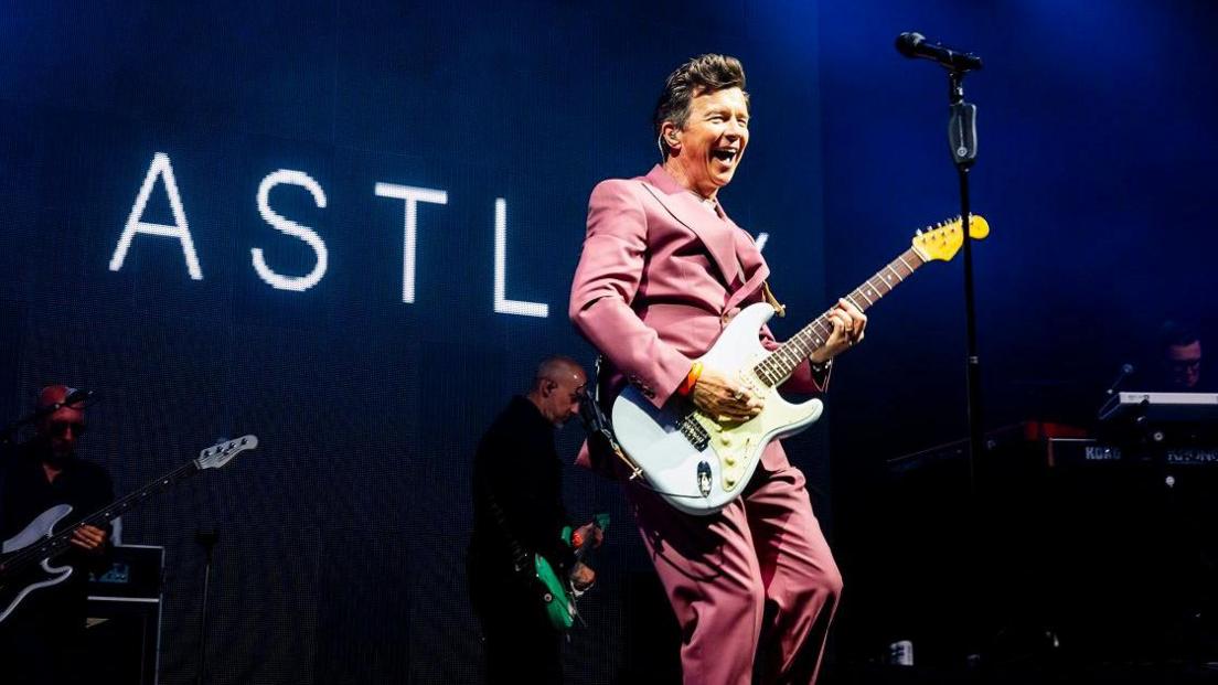 Rick Astley on stage at Bestival with a sign saying "Astley" behind him, he holds a guitar and wears a pink suit