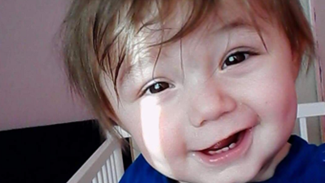 The image shows a child smiling at the camera standing in what appears to be a crib. The background includes a partially visible door and walls painted in light colours.