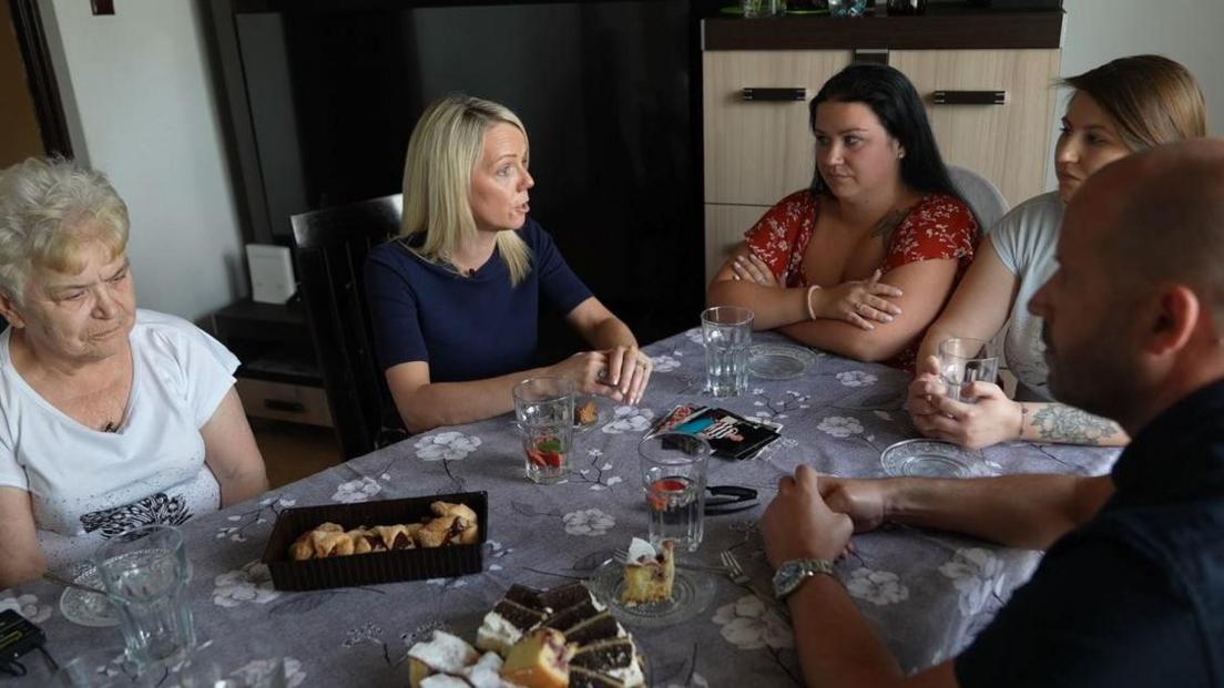 A male and female police officer sit at a kitchen table laden with cakes with the mother and two daughters of Monika