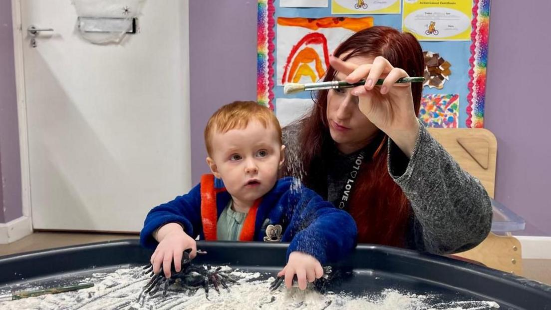 A small boy plays in with a woman in a children's play centre setting. In front of them is a large shallow sandpit filled with flour. The woman has long red hair and is holding a paintbrush sprinkling flour into the pit. She wears a greay cardigan and lanyard which says 'LOVE' repeatedly. The young boy is looking at the sprinkling flour whilst holding two toy spiders. He has short ginger hair and is wearing a blue dressing gown with a Mickey Mouse embroidered logo.