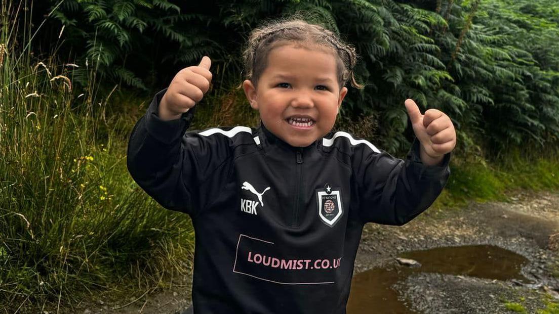 Karter with his thumbs up on the side of a hill with ferns behind him and a puddle on the footpath