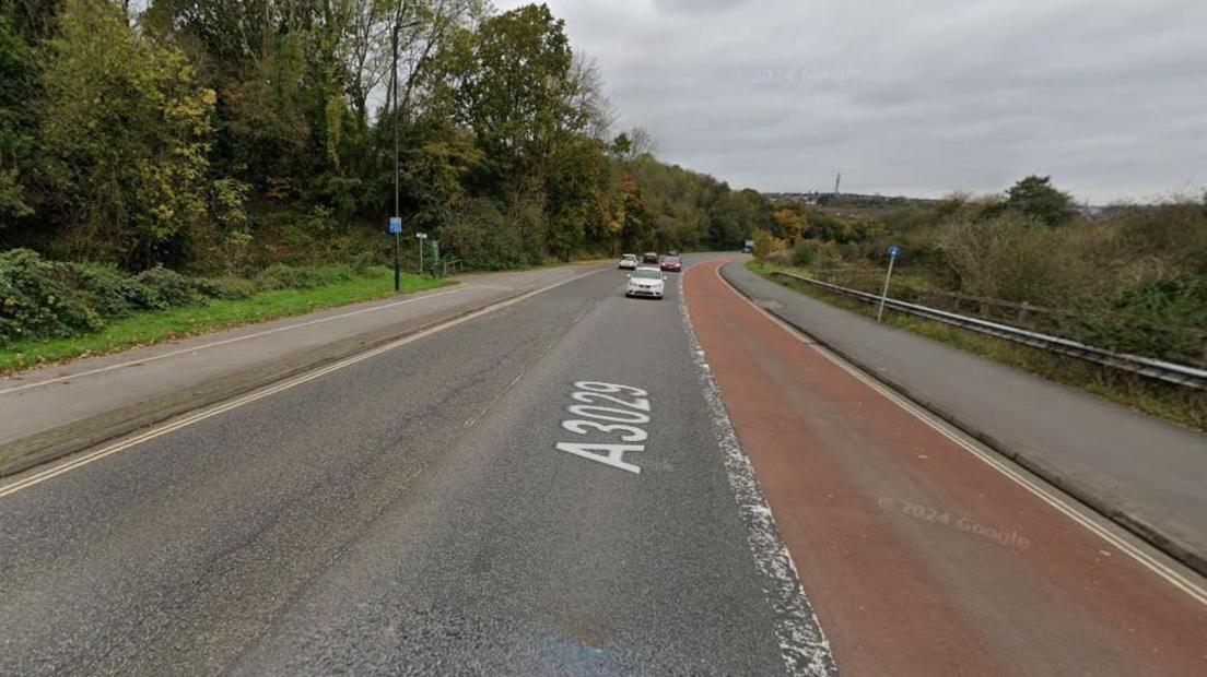 A shot looking down an A-road, cars coming towards the camera. Three lanes, one for buses. Greenery on either side, a path and cycle path on one also.