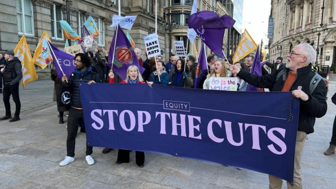 A group of protestors with banners and flags reading Stop The Cuts outside Birmingham City Council house