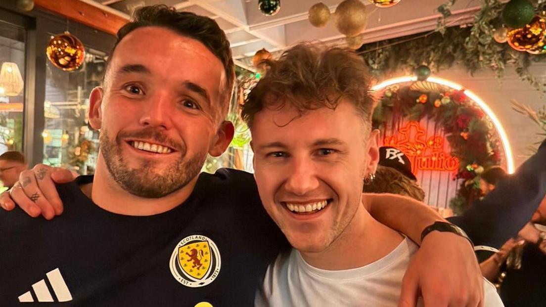 Scotland star John McGinn - in a Scotland team T-shirt, puts his arm around a beaming Callum Beattie in a busy Glasgow restaurant, decked out in Christmas decorations