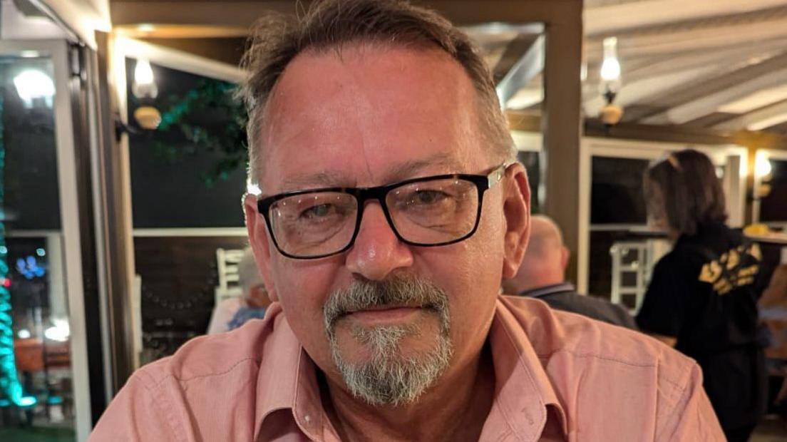 Spencer Clark sits in a restaurant while wearing a light pink shirt and large black-rimmed square glasses. He has a grey goatee beard.