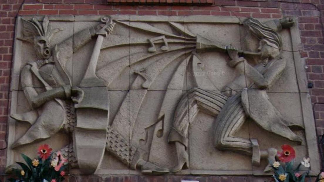 A concrete shallow relief sculpture in modernist style depicting two musicians playing instruments. The artwork is set into an old red brick wall.