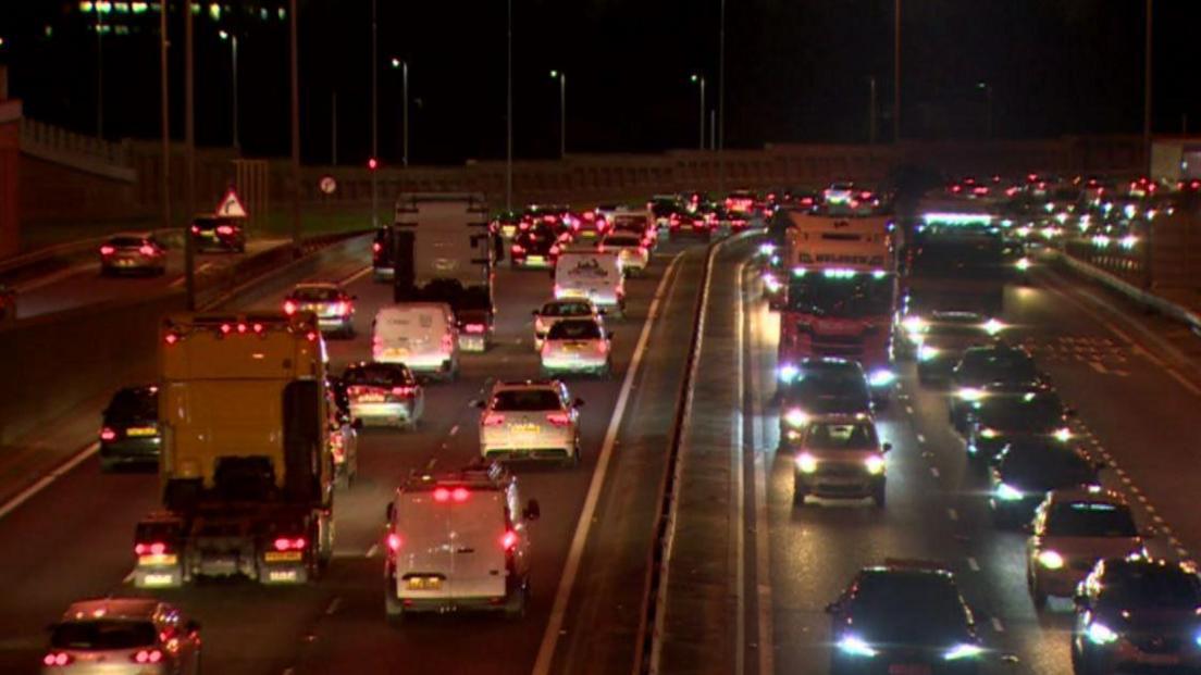 Cars, vans and lorries travelling along a motorway out of Belfast city centre. It is evening time and their headlights and taillights are on.