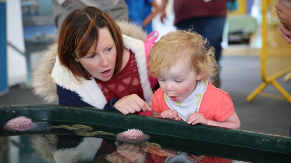 A little girl with curly blonde hair looks over into a tank with marine life, she looks wowed by what she can see. Her mum crouches next to her with a similar expression.