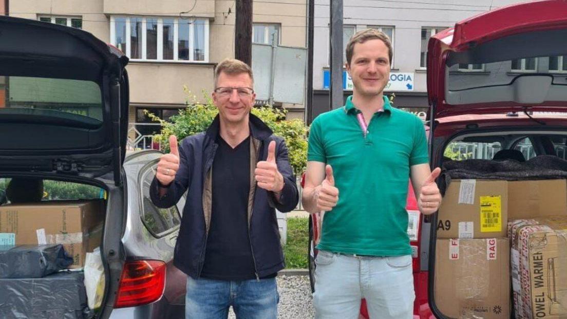 Mr Krivski stands with his thumbs up looking at the camera, next to a young man who also has his thumbs up. They pair are standing in front of two cars, both with their boots open containing cardboard boxes full of supplies. Mr Krivski is wearing thin wire framed glasses, a navy blue v neck t shirt with a blue jacket over the top and jeans. The man next to him has short dark blonde hair and wears a mint green polo top with pale denim jeans.