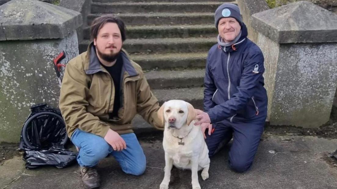 Zac Robinson with Ellie the cream labrador and James Elliott kneeling down at the bottom of stairs and looking at the camera with a litter picker behind them