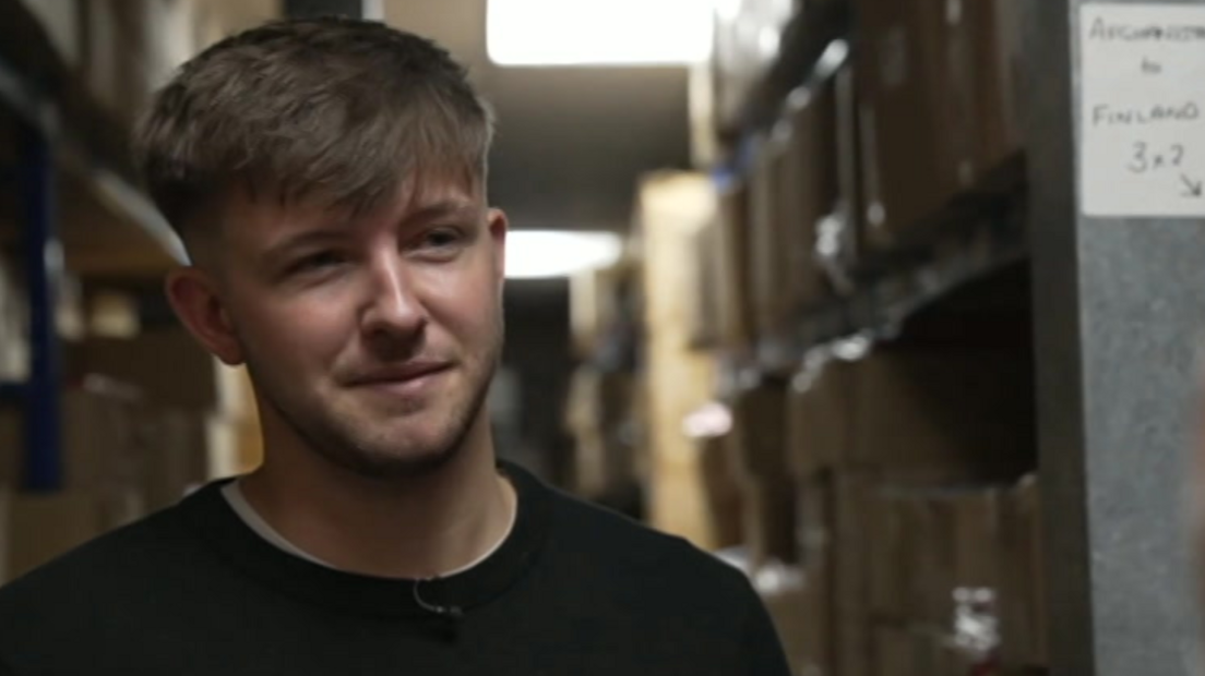 A man with dark hair, wearing a black jumper over a white t-shirt is looking across the camera, and smiling. Behind him is shelving with boxes on 