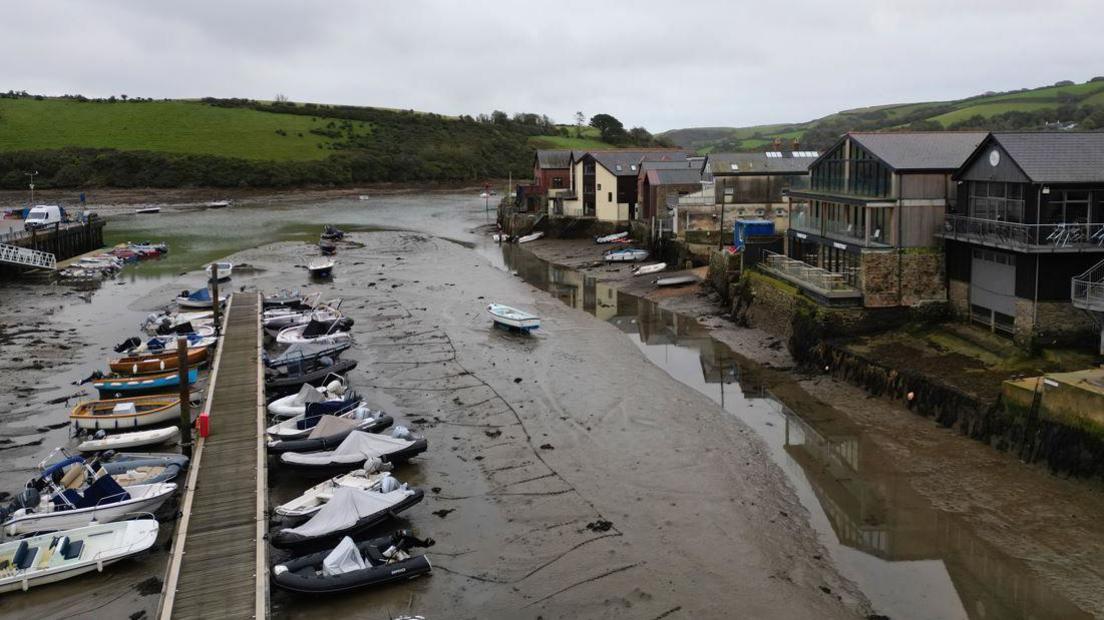 An inlet with a row of dinghies tied up along a pontoon on one side and a row of buildings, some industrial looking on the right.