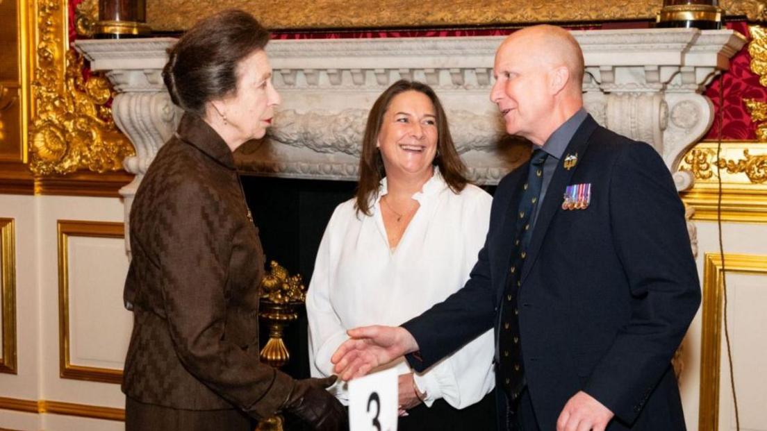 Andrew Gibson with the Princess Royal. He is wearing a suit with medals and is shaking her hand