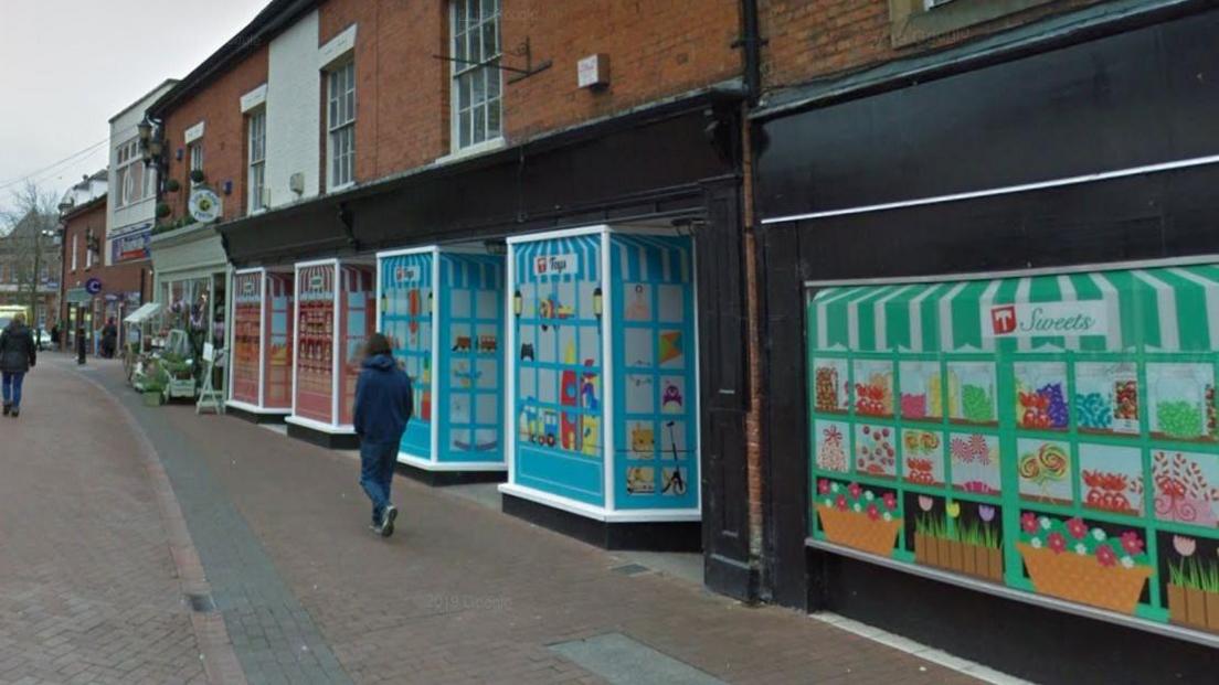A street view image of a town centre street. Terraced buildings with shop fronts are on the right. People can be seen walking past.
