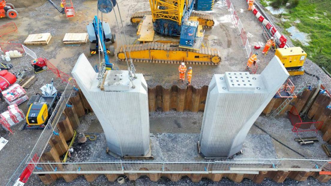 Two concrete piers surrounded by brown metal sheets. They have steel wires going upwards from them. Two workers in orange overalls are visible beyond the piers. Various pieces of building machinery are visible.