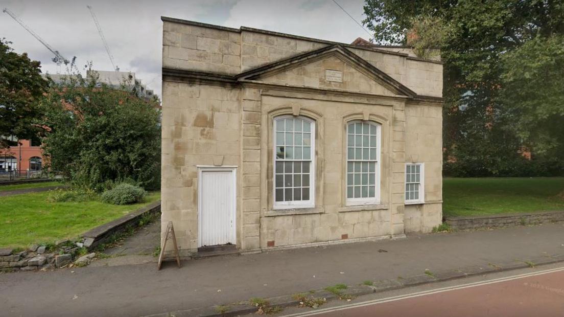 The front facade of Pile Street School in Redcliffe, right by the road. It is a limestone coloured building with two large windows at the front and a white door on the left that leads to nowhere.  