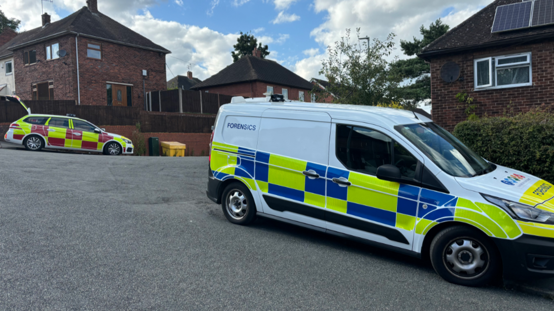 A fire service vehicle and a police forensics van parked outside properties on a residential street