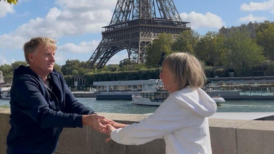 A man in a navy hoodie dances with a woman in a white hoodie by a river. The Eiffel Tower and several boats are in the background. 