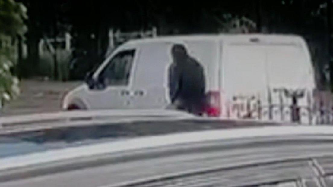 A man dressed in black with a black balaclava skulks next to a white van parked on the pavement in a residential street