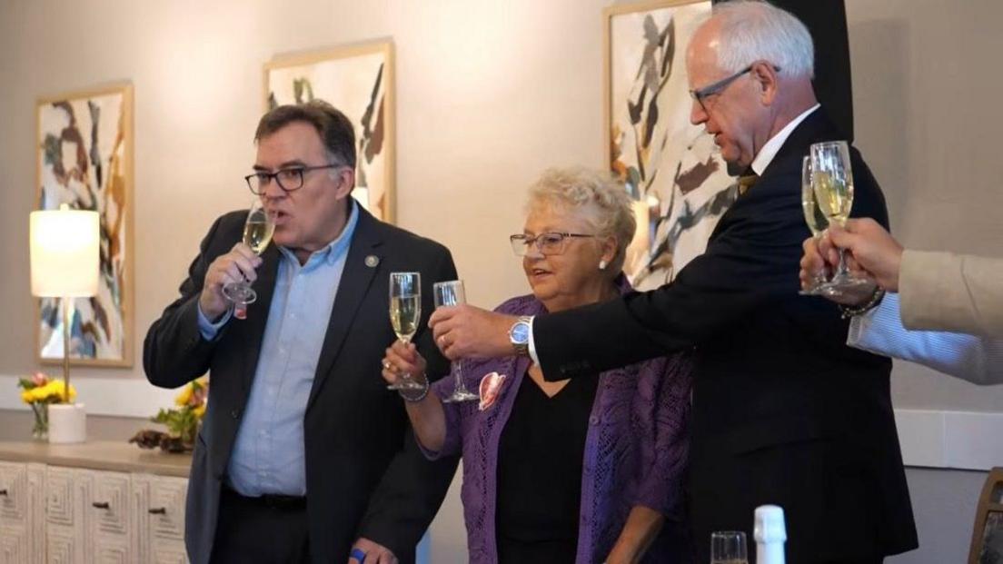 Anita Le Brun wears a purple blazer and glasses as she holds up a flute glass of champagne to toast Minnesota Gov Tim Walz, who is taller, balding and holding his own glass close to hers