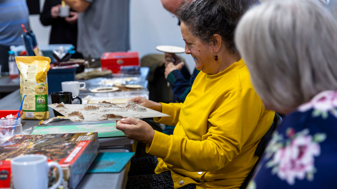 A woman in a yellow jumper hold something she is making in her hands. She has model making kit and paraphernalia on a table in front of her.A lady wit grey hair and a patterned top is sat in the foreground wit others in the background.