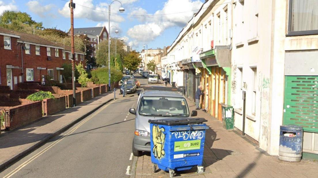 An image from Google Maps Street View of Grosvenor Road in St Pauls, Bristol. The photograph is taken on a sunny day and there are cars parked either side of the street and a large blue industrial wheelie bin in the foreground