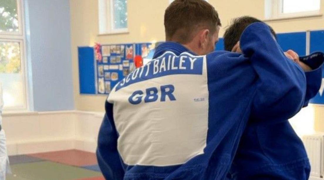 Scott Bailey with his back to the camera in judo action with someone else. Blue boards are on the wall behind them below windows.
