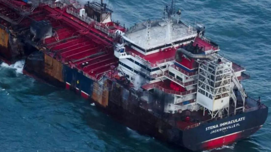 A large oil tanker with a gaping hole in its port [left] side. The area around the bridge, at the stern of the ship, is blackened from fire. 